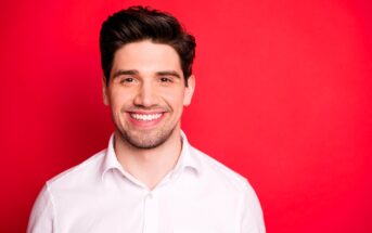 Smiling man with dark hair wearing a white shirt stands in front of a bright red background.