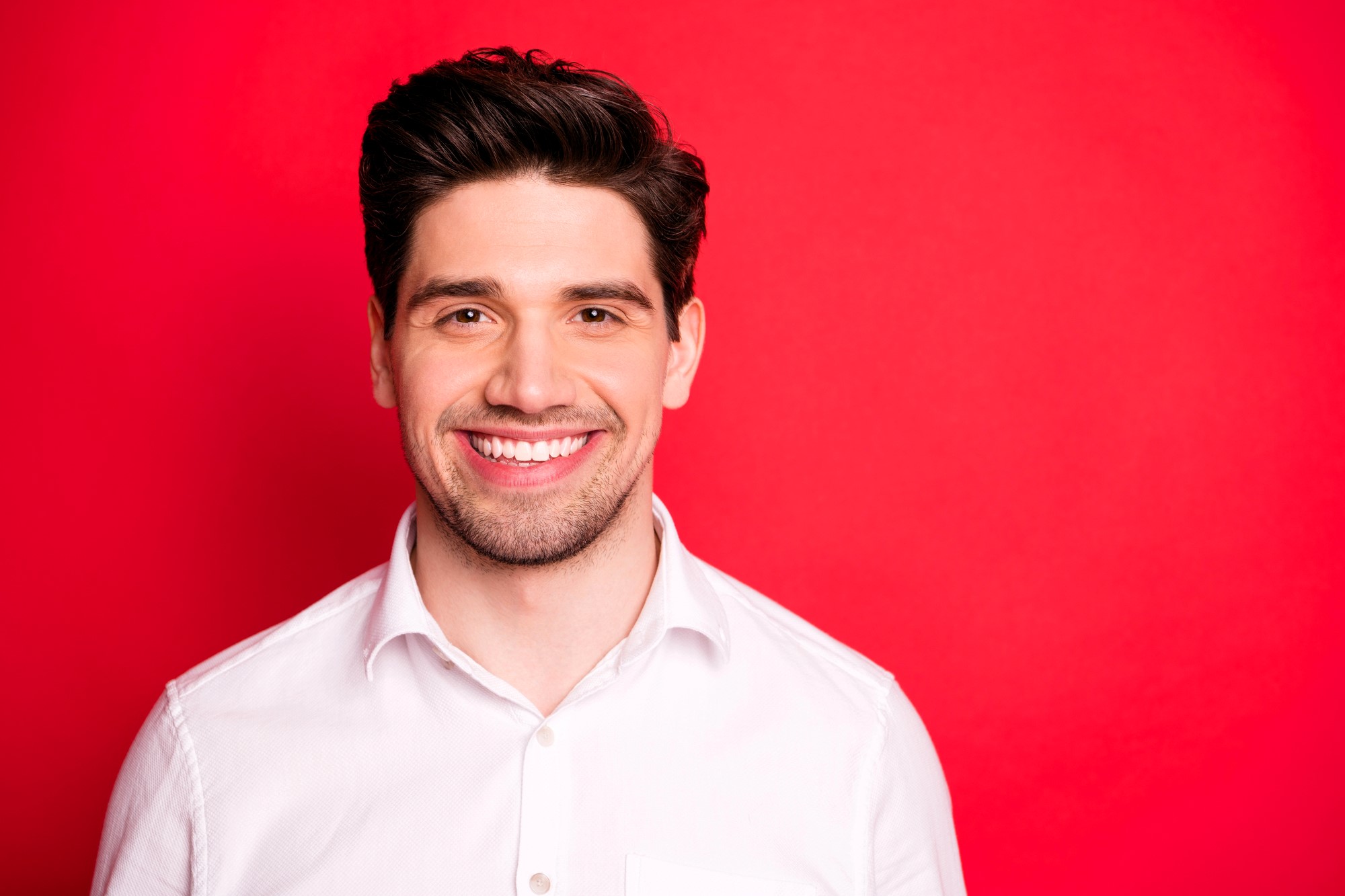 Smiling man with dark hair wearing a white shirt stands in front of a bright red background.