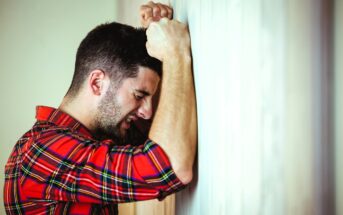 A man in a red plaid shirt leans his forehead against a wall, appearing distressed or frustrated, with his eyes closed and hands pressed against the wall.