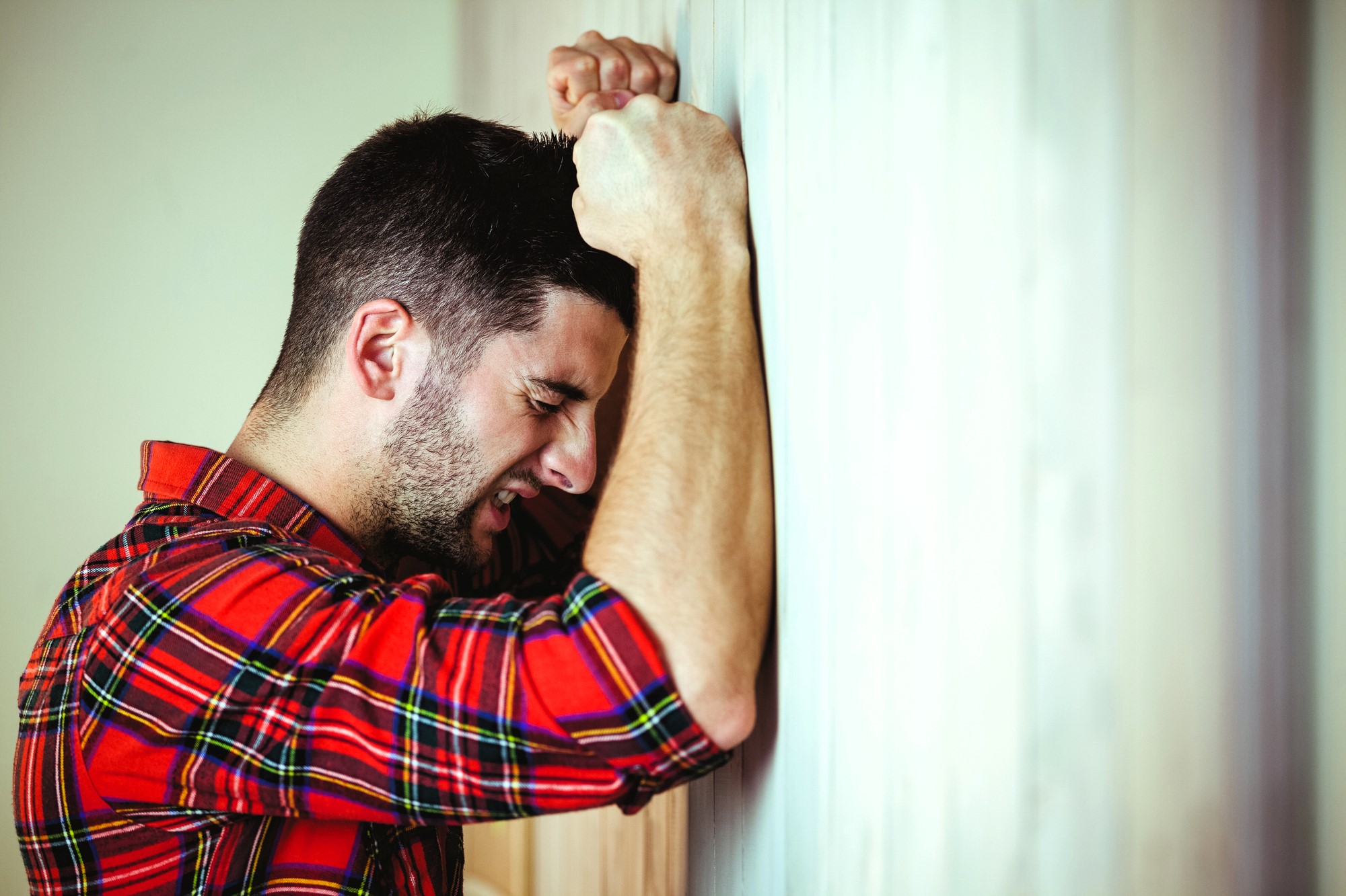 A man in a red plaid shirt leans his forehead against a wall, appearing distressed or frustrated, with his eyes closed and hands pressed against the wall.