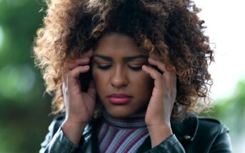 Person with curly hair holds their temples, eyes closed, appearing to concentrate or feel discomfort. They wear a striped top and leather jacket, standing outdoors with blurred greenery in the background.