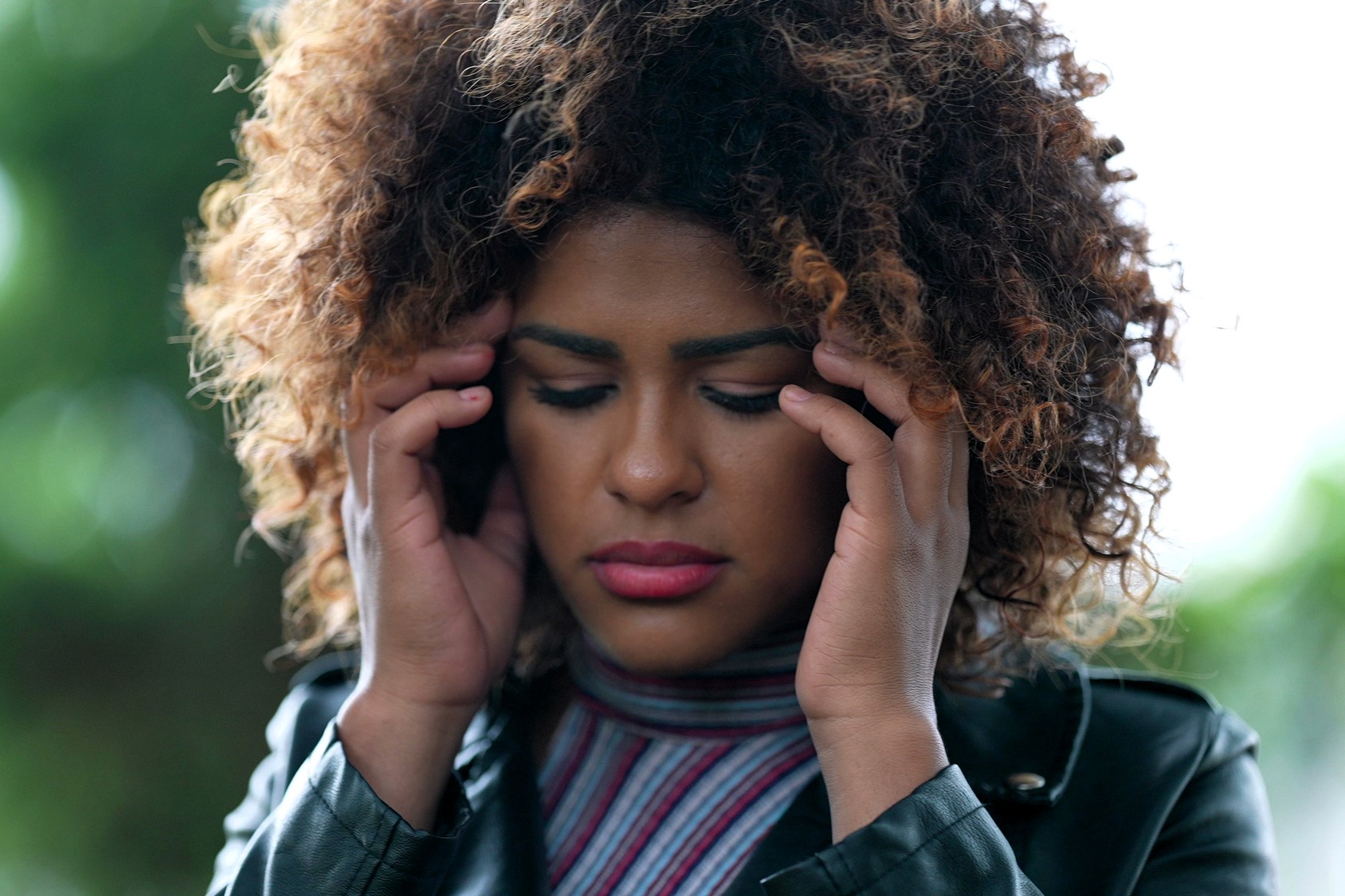 Person with curly hair holds their temples, eyes closed, appearing to concentrate or feel discomfort. They wear a striped top and leather jacket, standing outdoors with blurred greenery in the background.