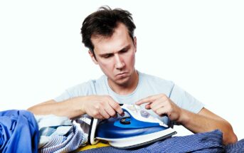 A man with brown hair looks intently at a blue and white iron in his hands. He is surrounded by a pile of clothes. He wears a light blue shirt and appears focused on the iron's settings or buttons.