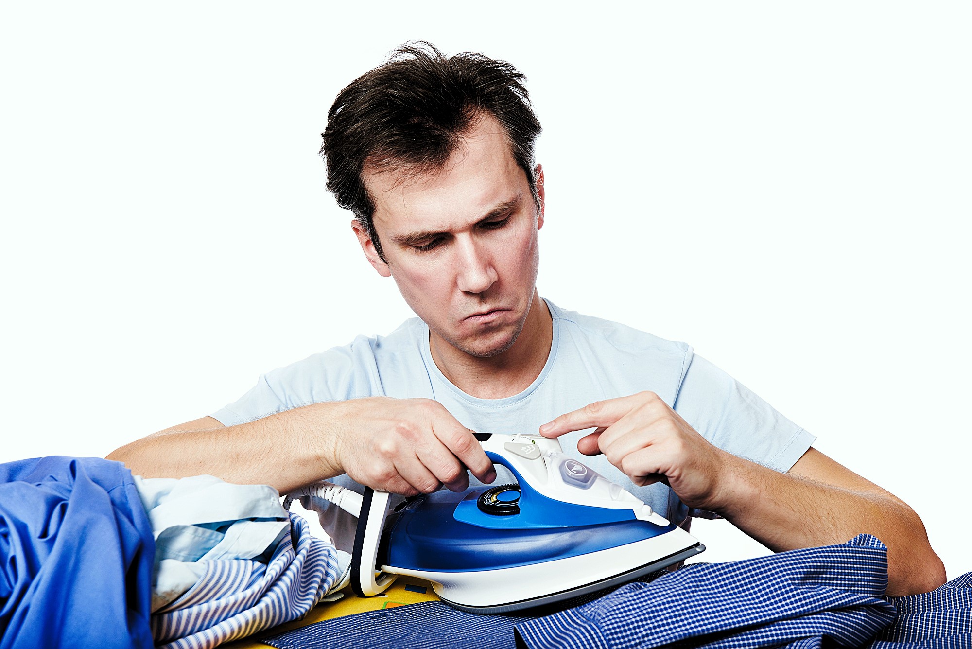 A man with brown hair looks intently at a blue and white iron in his hands. He is surrounded by a pile of clothes. He wears a light blue shirt and appears focused on the iron's settings or buttons.