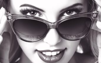 Black and white close-up of a person wearing cat-eye sunglasses, smiling slightly with visible teeth. Their hands adjust the frames, and their eyes are partially hidden behind the lenses.