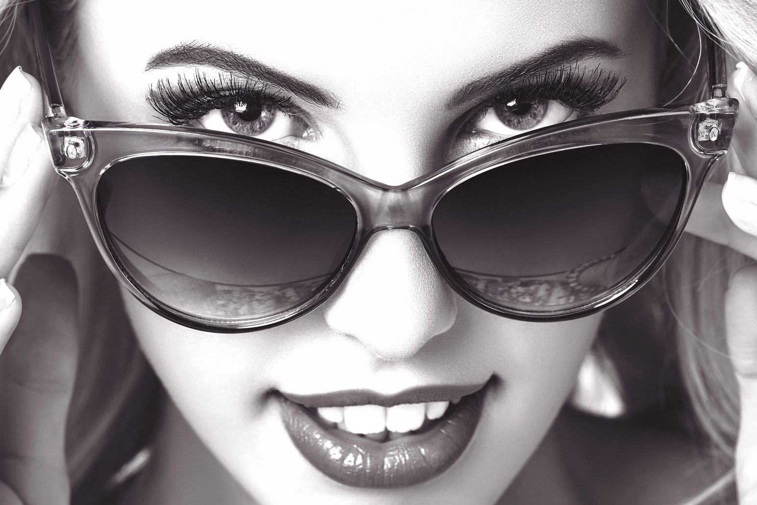 Black and white close-up of a person wearing cat-eye sunglasses, smiling slightly with visible teeth. Their hands adjust the frames, and their eyes are partially hidden behind the lenses.