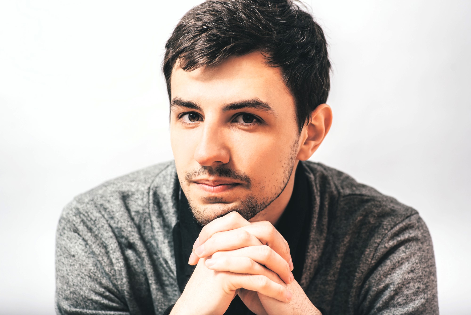 A man with short dark hair and a beard is looking at the camera with a slight smile. He is wearing a dark gray sweater and is resting his chin on his interlocked hands against a light gray background.