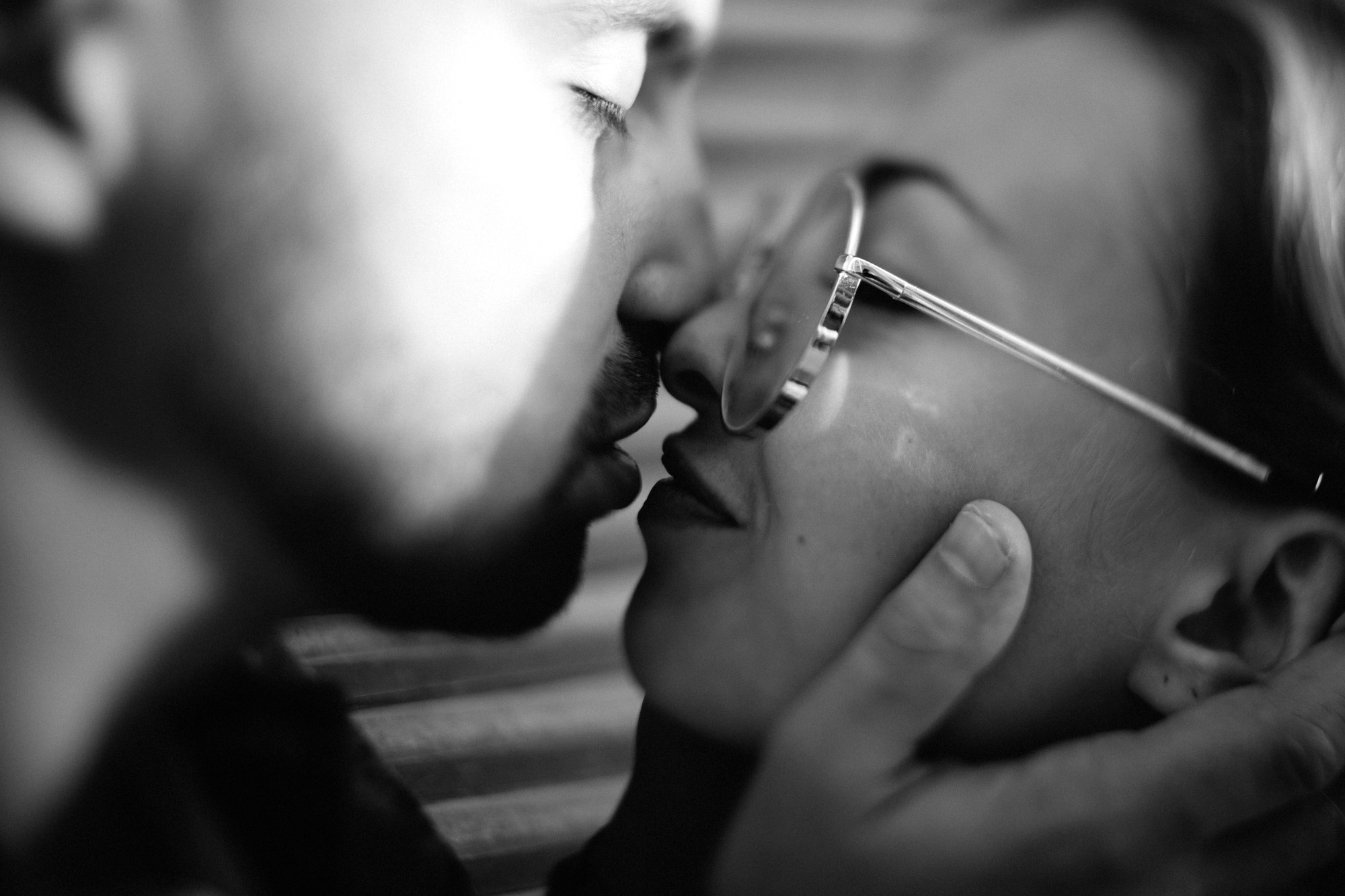 A black and white close-up of a couple about to kiss. The man has facial hair and gently holds the woman's face. The woman is wearing glasses, and both are lit by soft light, creating an intimate atmosphere.