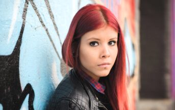 A person with long red hair stands in front of a graffiti-covered wall. They are wearing a black leather jacket and a plaid shirt, looking directly at the camera with a neutral expression.