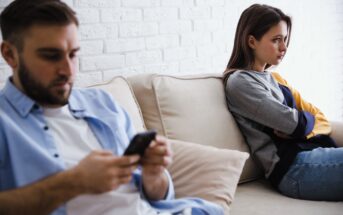 A man and a woman sit on a beige couch. The man is in the foreground, looking at his phone. The woman is in the background, arms crossed, looking away, appearing upset. They are against a white brick wall.