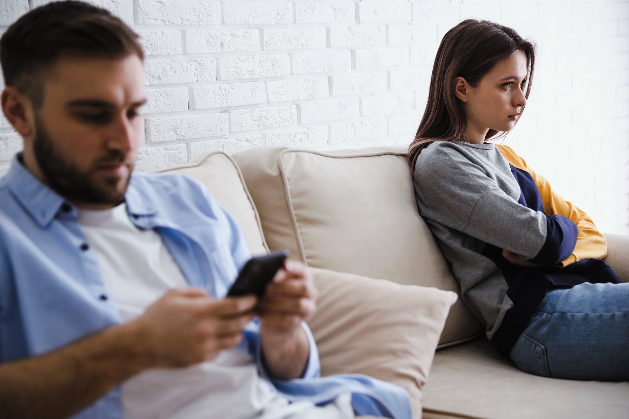 A man and a woman sit on a beige couch. The man is in the foreground, looking at his phone. The woman is in the background, arms crossed, looking away, appearing upset. They are against a white brick wall.