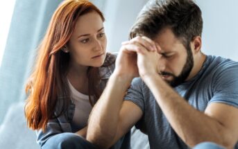 A woman with red hair looks concerned and supportive as she sits next to a man with brown hair, who appears upset and is resting his head on his hands. They are indoors, and he seems to be experiencing distress.