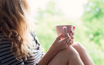 A woman with wavy blonde hair holding a pink mug sits on a ledge, wearing a black and white striped shirt. She is looking out at a blurred, leafy green background, suggesting a peaceful outdoor scene.