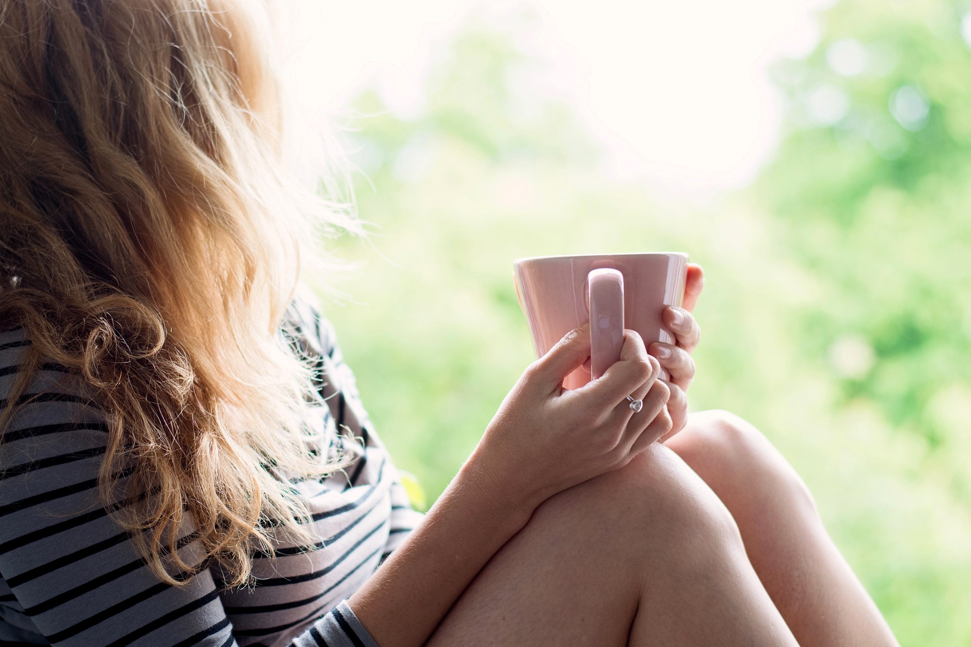 A woman with wavy blonde hair holding a pink mug sits on a ledge, wearing a black and white striped shirt. She is looking out at a blurred, leafy green background, suggesting a peaceful outdoor scene.