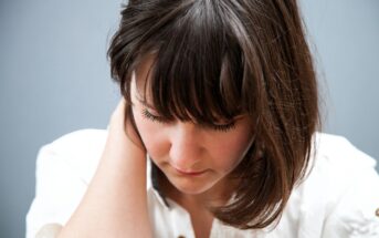 Woman with shoulder-length brown hair looks down, gently touching the back of her neck with one hand. She wears a white shirt with buttons and appears to be in a serene or contemplative mood against a plain gray background.