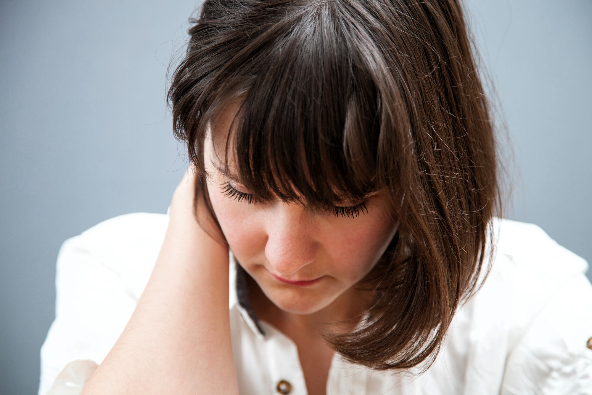 Woman with shoulder-length brown hair looks down, gently touching the back of her neck with one hand. She wears a white shirt with buttons and appears to be in a serene or contemplative mood against a plain gray background.