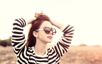 Woman in a striped sweater wearing sunglasses, standing outdoors with hands in her hair. The background features a soft, blurred landscape and a warm, sunlit ambiance.