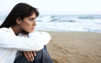 A woman in a white shirt and jeans sits on a sandy beach, gazing thoughtfully towards the sea. The ocean waves are gently rolling in under a cloudy sky. Her long dark hair is flowing freely in the breeze.