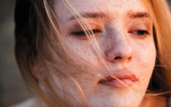 Close-up of a person with freckled skin and light hair, partially obscured by strands of hair blowing in the wind. Their eyes are softly closed or looking downwards, creating a serene and thoughtful expression.