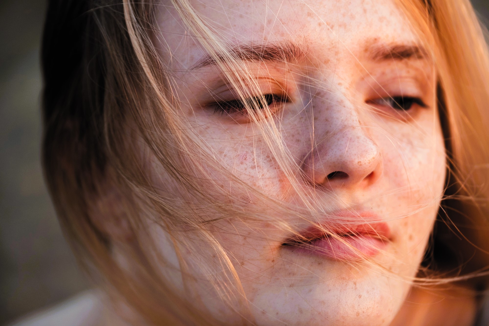 Close-up of a person with freckled skin and light hair, partially obscured by strands of hair blowing in the wind. Their eyes are softly closed or looking downwards, creating a serene and thoughtful expression.