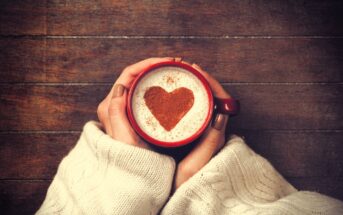 Hands in a cozy sweater hold a red mug of frothy coffee with a heart shape made of cocoa on top, set against a wooden background.