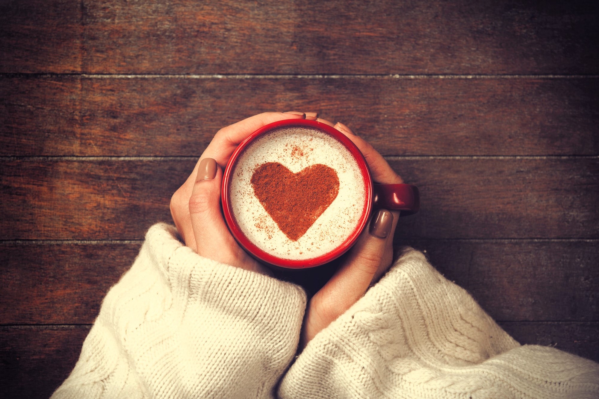 Hands in a cozy sweater hold a red mug of frothy coffee with a heart shape made of cocoa on top, set against a wooden background.