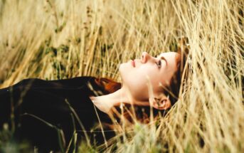 A person with long hair lies in a field of tall, dry grass, gazing upward. They are wearing a dark shirt, and the scene conveys a sense of tranquility and contemplation amidst nature.