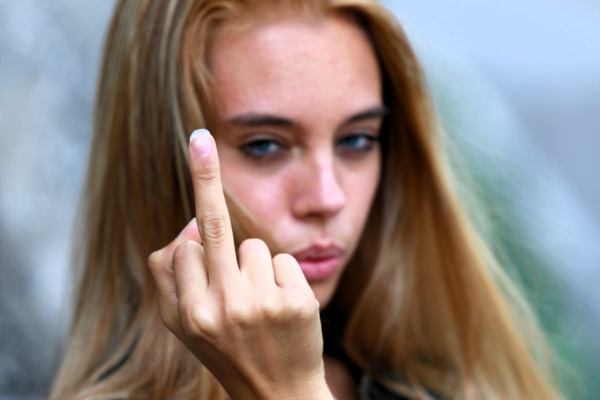 A person with long hair holds up their middle finger, partially blurred background.