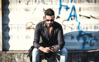 A man with a beard and sunglasses sits on a concrete ledge, looking at his phone. He is wearing a black shirt and light blue jeans. The background has a corrugated metal wall with graffiti.