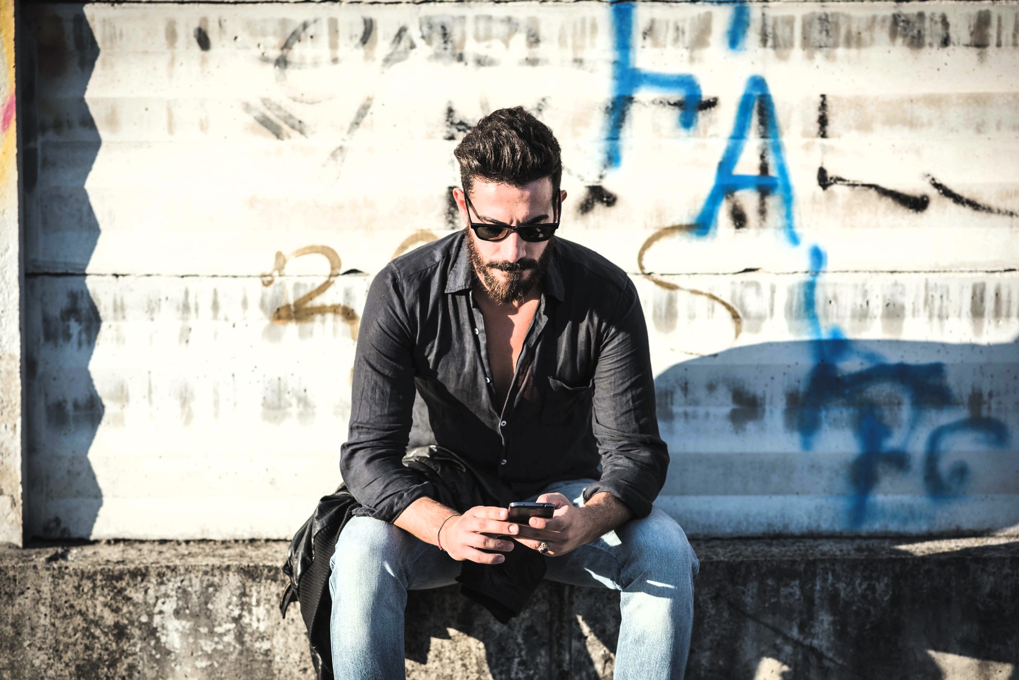 A man with a beard and sunglasses sits on a concrete ledge, looking at his phone. He is wearing a black shirt and light blue jeans. The background has a corrugated metal wall with graffiti.