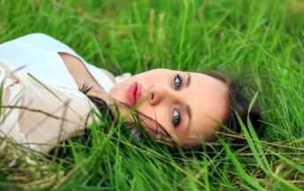 Woman with dark hair and blue eyes lying on her back in lush green grass, looking at the camera. She is wearing a light-colored top, and the background is filled with vibrant grass blades.