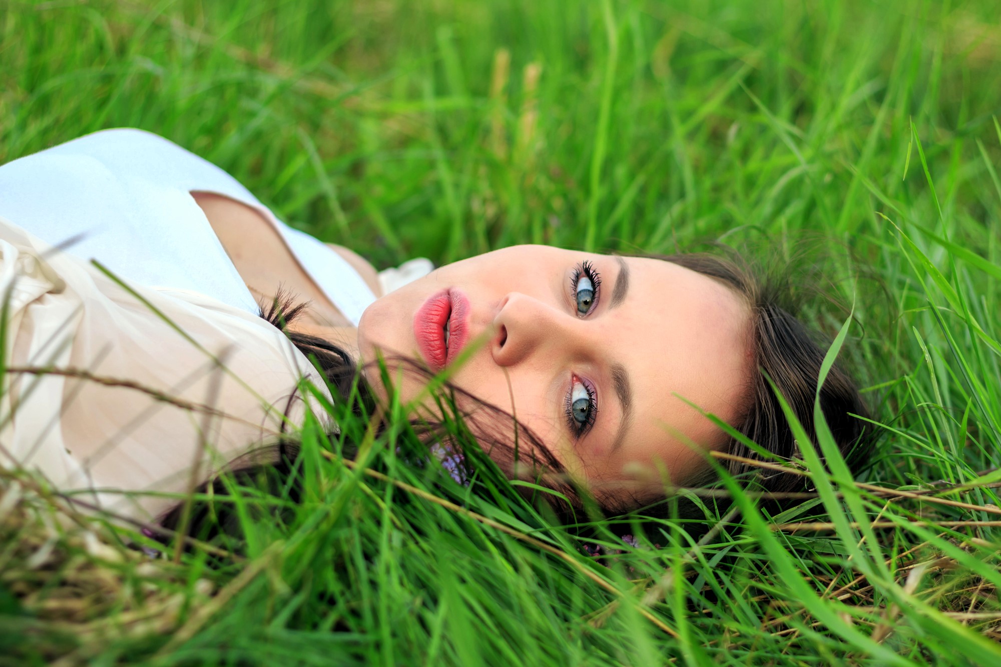 Woman with dark hair and blue eyes lying on her back in lush green grass, looking at the camera. She is wearing a light-colored top, and the background is filled with vibrant grass blades.