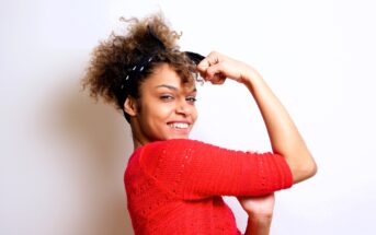 Woman in a red sweater smiling confidently, with her arm raised and flexed, reminiscent of the "We Can Do It" pose. Her hair is curly, and she wears a black headband, standing against a plain white background.