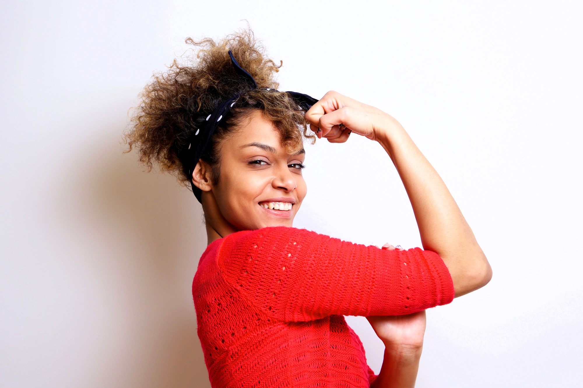 Woman in a red sweater smiling confidently, with her arm raised and flexed, reminiscent of the "We Can Do It" pose. Her hair is curly, and she wears a black headband, standing against a plain white background.