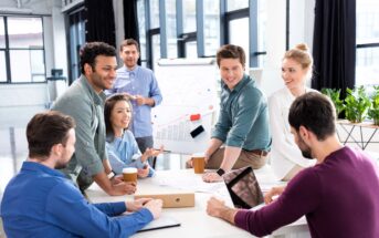 A group of people, three men and three women, are smiling and collaborating in a modern office setting. One person is using a laptop, while others reference a presentation with charts on a whiteboard. Coffee cups and a box are on the table.