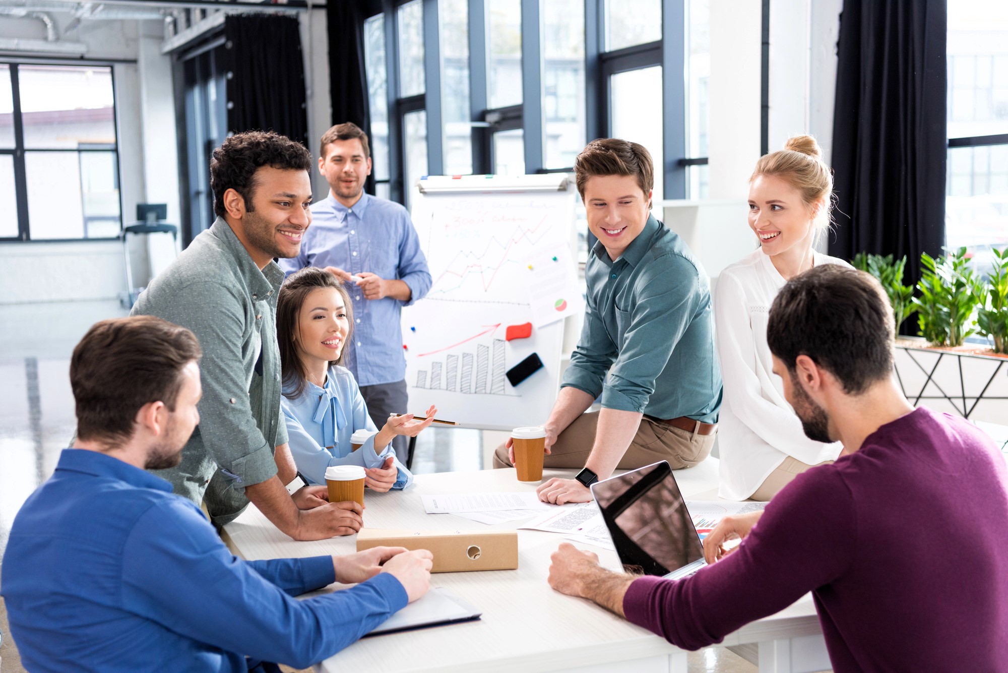 A group of people, three men and three women, are smiling and collaborating in a modern office setting. One person is using a laptop, while others reference a presentation with charts on a whiteboard. Coffee cups and a box are on the table.