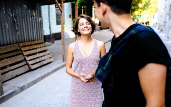 A woman in a light dress smiles while looking at a person with short dark hair, wearing a black shirt and holding a plaid item. They are standing on a street with construction materials and trees in the background.