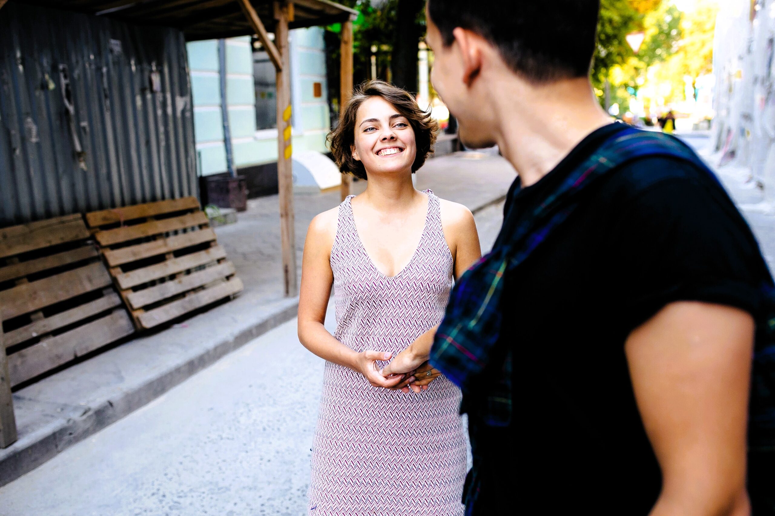 A woman in a light dress smiles while looking at a person with short dark hair, wearing a black shirt and holding a plaid item. They are standing on a street with construction materials and trees in the background.