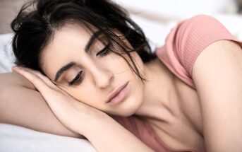 A woman with dark hair lies on her side, resting her head on her arm. She has a neutral expression, closed eyes, and wears a pink top. The background is softly blurred, emphasizing her serene pose.