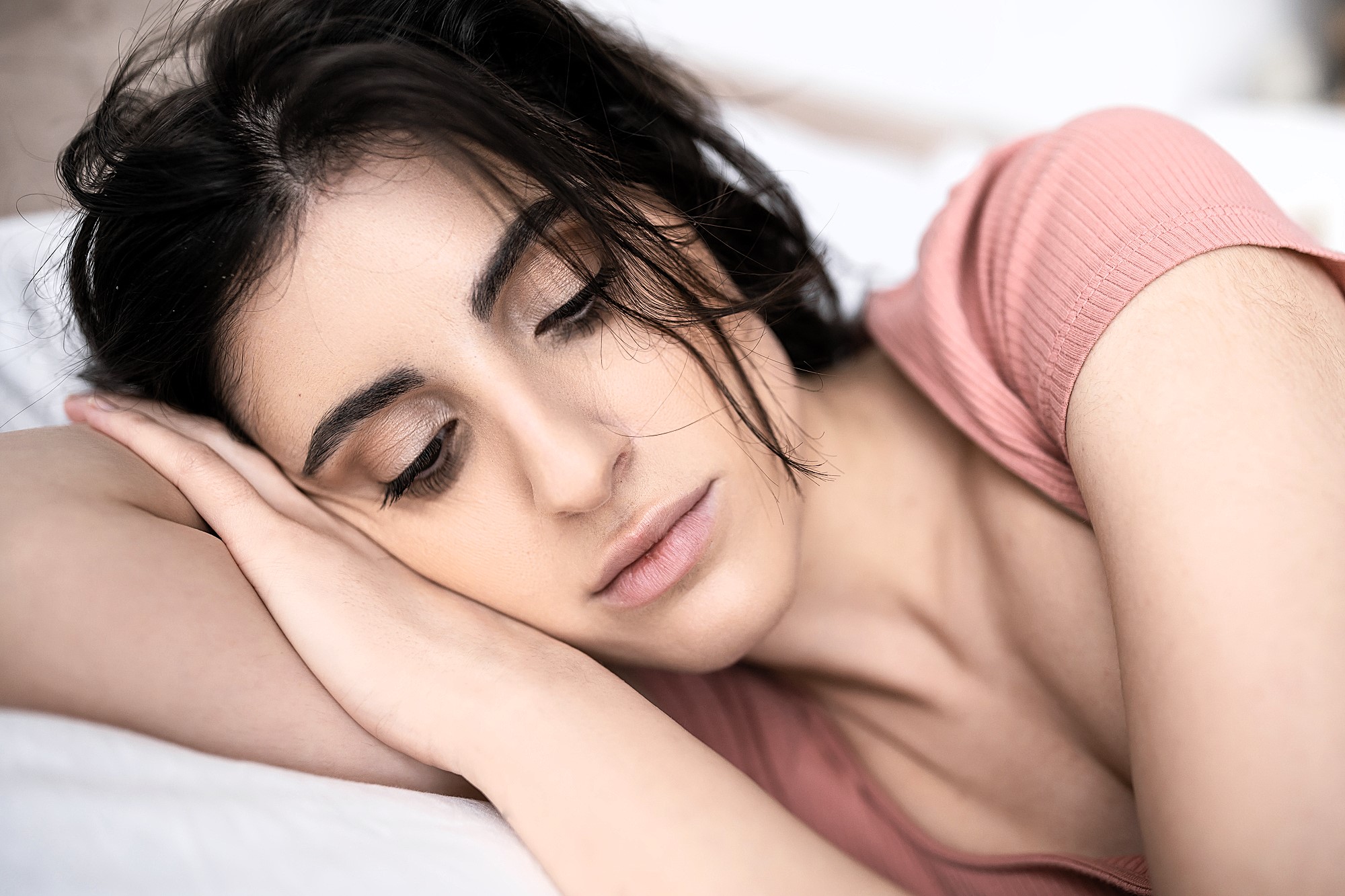 A woman with dark hair lies on her side, resting her head on her arm. She has a neutral expression, closed eyes, and wears a pink top. The background is softly blurred, emphasizing her serene pose.