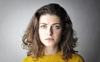 Woman with medium-length brown hair and green eyes, wearing a mustard yellow sweater, stares seriously at the camera against a neutral gray background.