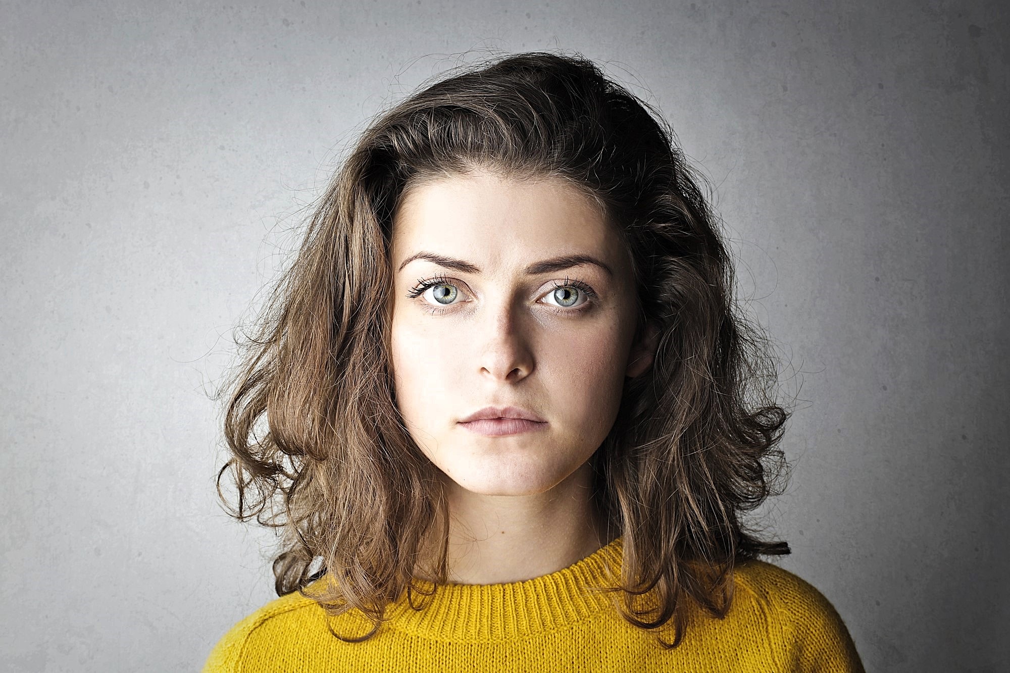 Woman with medium-length brown hair and green eyes, wearing a mustard yellow sweater, stares seriously at the camera against a neutral gray background.