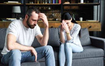 A man and woman sit on a couch, appearing upset. The man rests his head in his hand, while the woman has her hands clasped near her face. Shelves with books and decorations are in the background.