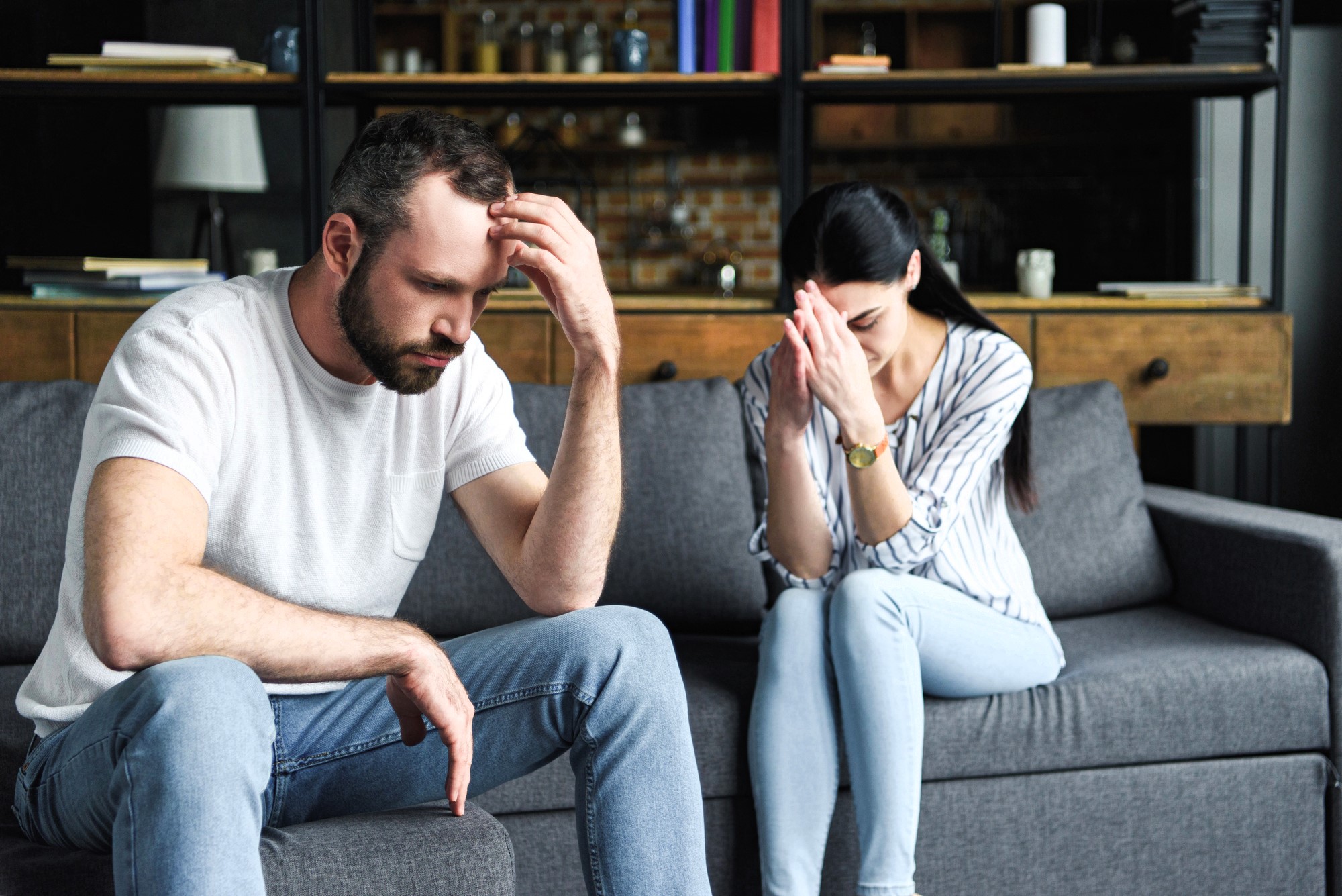 A man and woman sit on a couch, appearing upset. The man rests his head in his hand, while the woman has her hands clasped near her face. Shelves with books and decorations are in the background.