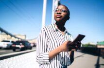 Person in a striped shirt and glasses stands on a bridge, holding a smartphone. They look upward with a thoughtful expression. The background shows a clear blue sky, a suspension wire, and blurred cars on the road.