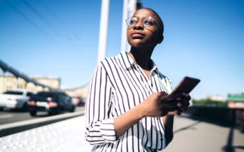 Person in a striped shirt and glasses stands on a bridge, holding a smartphone. They look upward with a thoughtful expression. The background shows a clear blue sky, a suspension wire, and blurred cars on the road.