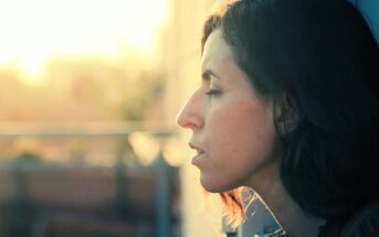 Side profile of a woman with shoulder-length dark hair, eyes closed, basking in the warm glow of sunlight. The background is softly blurred, giving a tranquil, contemplative atmosphere.