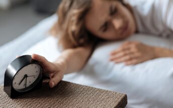 A person lying in bed reaches out to turn off a black alarm clock on a wicker nightstand. The time on the clock reads 7:00. The background is softly blurred, conveying a sleepy, early morning atmosphere.