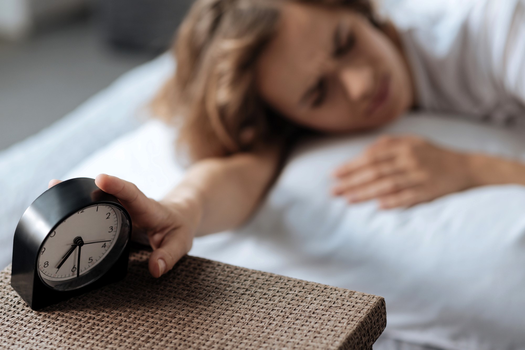 A person lying in bed reaches out to turn off a black alarm clock on a wicker nightstand. The time on the clock reads 7:00. The background is softly blurred, conveying a sleepy, early morning atmosphere.