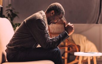 A man sitting on a sofa, leaning forward with his elbows on his knees and holding a smartphone to his forehead. He appears deep in thought or concerned. The room is softly lit with neutral-toned furnishings in the background.